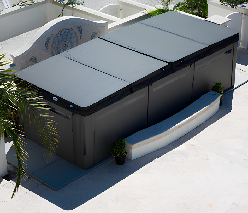 A large outdoor hot tub with a dark gray cover and black trim, photographed on a sunny patio with plants and white walls in the background.